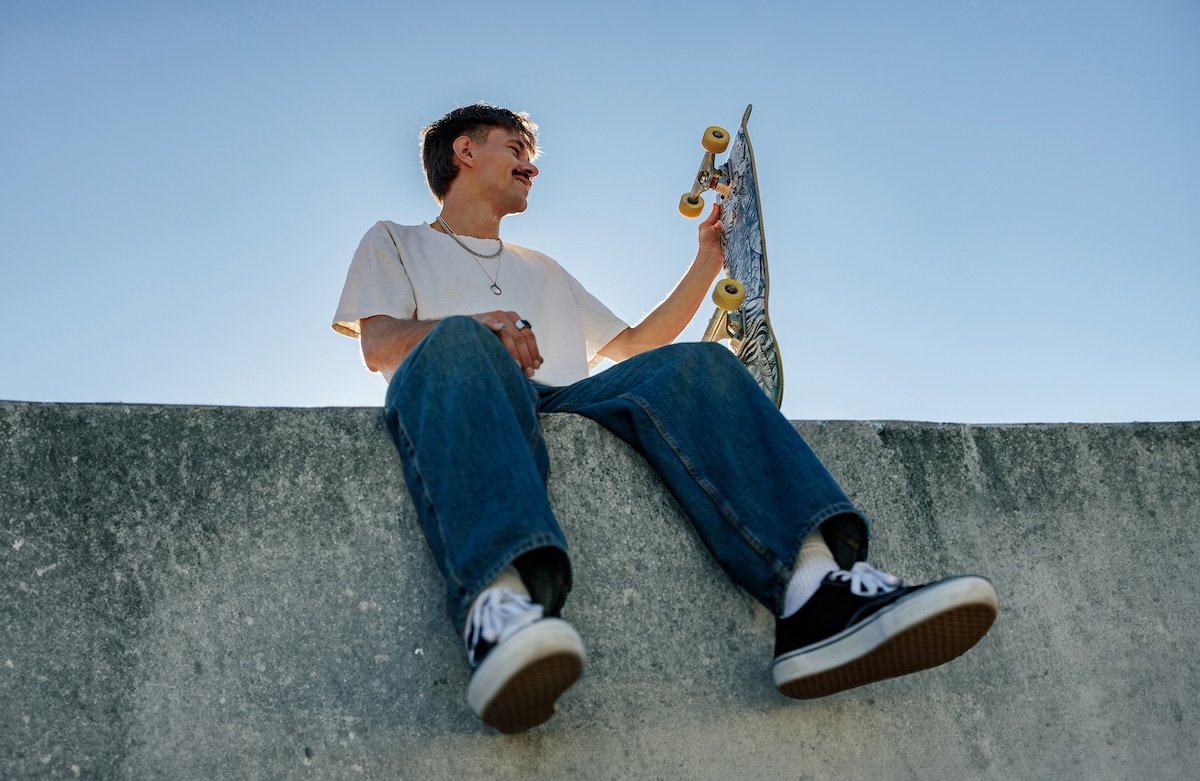 Low angle of young male skater in casual outfit sitting on ramp with skateboard against blue sky