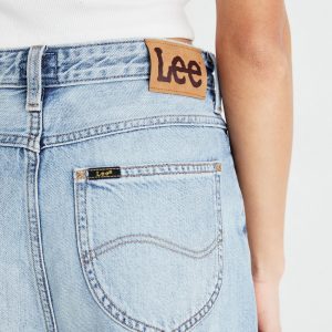 Close-up of a person wearing light-wash denim jeans, showing a tan Lee patch on the waistband and back pocket stitching.