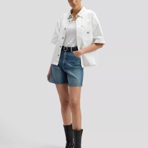 Female model wearing a white short-sleeve button-up shirt over a white tank top, blue denim shorts, a black belt, and black ankle boots.