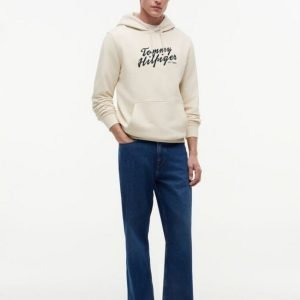 Male model wearing a cream Tommy Hilfiger hoodie with blue jeans and brown shoes, facing forward.