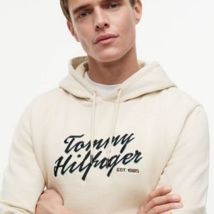 Male model wearing a cream hoodie with a bold Tommy Hilfiger logo on the chest, arms crossed in a studio setting