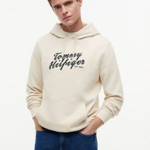 Model wearing a cream Tommy Hilfiger hoodie with EST. 1985 logo, standing with hands in pockets against a light background.