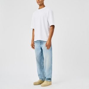 Full-body shot of a man in a white oversized T-shirt, light blue baggy jeans, and beige loafers against a plain light background.