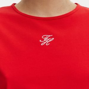 Red shirt with a small white cursive monogram logo near the neckline, close-up detail.