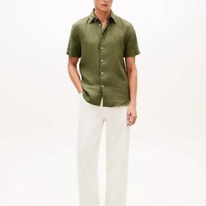 Male model wearing an olive green short-sleeve button-down shirt with white trousers and black loafers, posing against a white background with hands in pockets.
