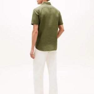 Man seen from behind wearing an olive short-sleeve shirt and white pants, standing in a studio against a pale background.