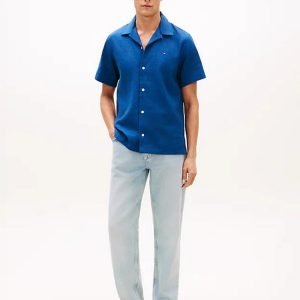 Young man wearing a cobalt blue short-sleeve button-down shirt, light-washed jeans, and beige shoes, standing with hands in pockets.