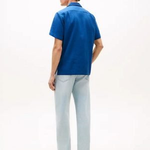 Man seen from behind wearing a blue short-sleeve shirt, light-wash jeans, and beige sneakers against a white background.