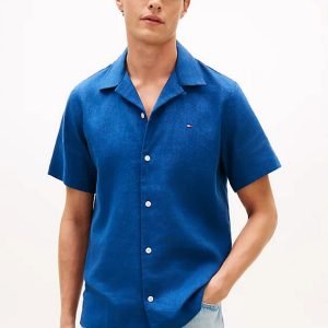 Model wearing a bright blue short-sleeve button-down shirt and light jeans, standing with one hand in pocket.