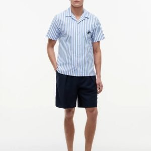 Young man in a light-blue and white striped short-sleeve shirt with navy shorts, standing with hands in pockets against a white background.