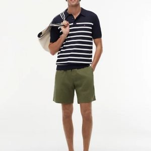 Man standing in a navy striped polo and olive shorts, holding a light tote over his shoulder, wearing a cap and beige loafers.