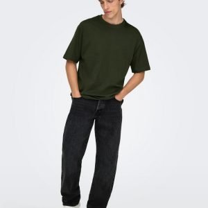 Young man posing in an olive oversized T-shirt, black jeans, and white sneakers with hands in pockets.