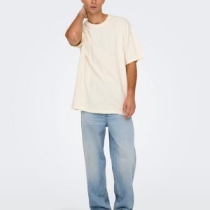 Man wearing an oversized cream T-shirt, light blue jeans, and cream sneakers, posing with one hand at his neck.