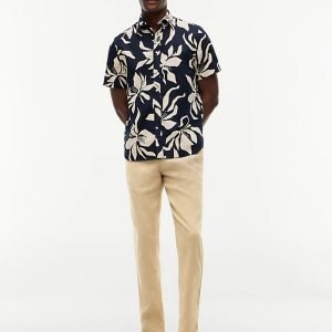 Man wearing a navy tropical print shirt, beige pants, and sandals with sunglasses, standing casually against a light background.