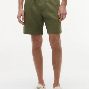 Man wearing olive green drawstring shorts with a striped navy and white shirt, standing in beige loafers against a white background.