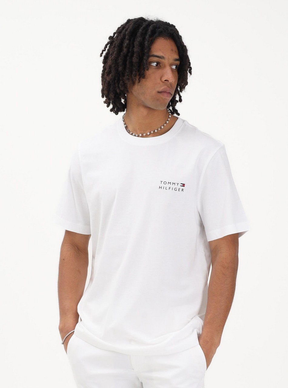 Young man in a white Tommy Hilfiger t-shirt with a small logo on the chest and a beaded necklace, posing against a white backdrop.