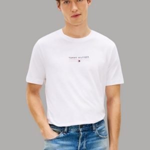 Young male model wearing a white Tommy Hilfiger logo tee and blue jeans, hands in pockets, gray background.