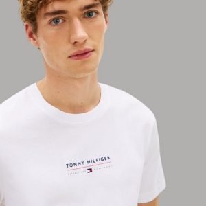 Young man with curly light brown hair wearing a white Tommy Hilfiger T-shirt against a gray background, looking slightly to the side.
