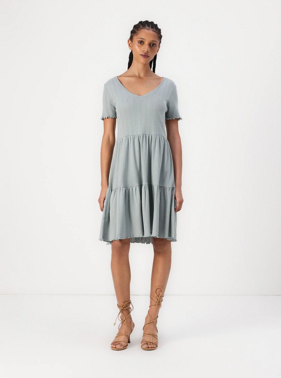 Woman wearing a light blue, short-sleeve, tiered dress with a V-neck, standing against a white background with tan lace-up sandals.