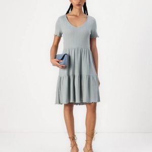 Woman in a light blue V-neck ribbed dress with a tiered, ruffled skirt, holding a small blue clutch and wearing tan lace-up sandals.