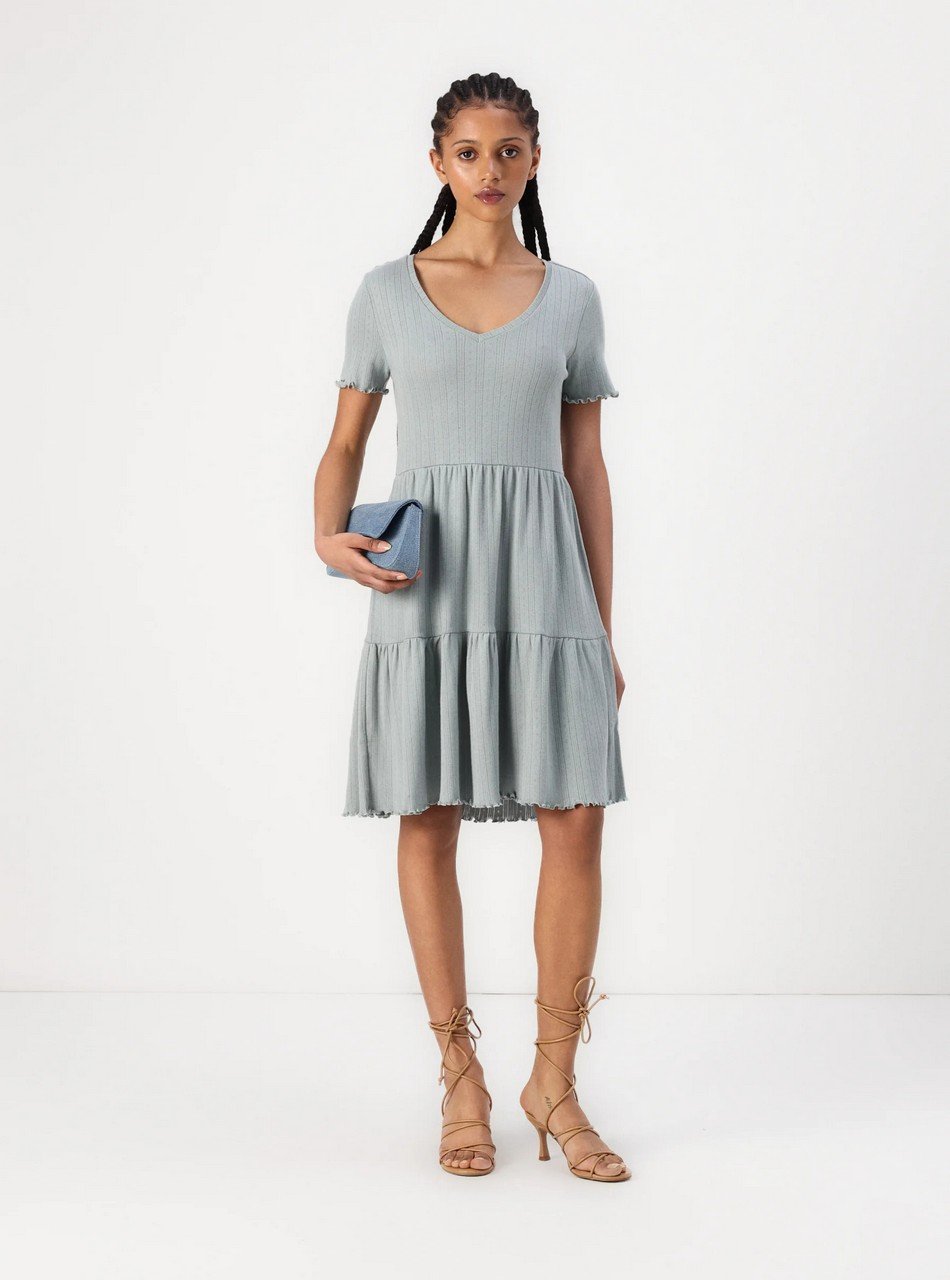 Woman in a light blue V-neck ribbed dress with a tiered, ruffled skirt, holding a small blue clutch and wearing tan lace-up sandals.