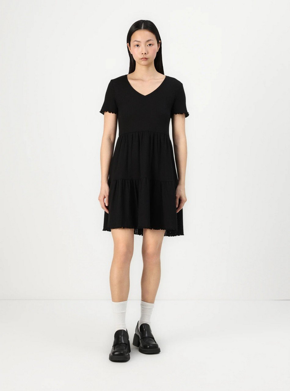Model wearing a black V-neck short-sleeve tiered dress with white socks and black loafers, standing against a white background.