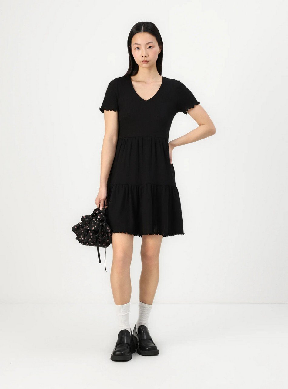 Woman modeling a black V-neck, short-sleeve tiered dress, white socks, black loafers, and a small black floral handbag against a white background.