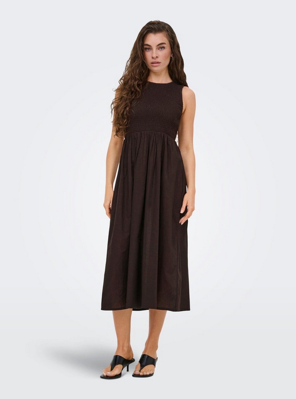 Woman wearing a sleeveless dark brown smocked-bodice midi dress with a full, flowing skirt and black sandals, standing facing forward.