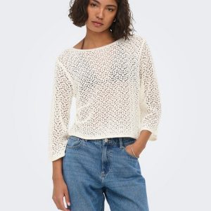 Woman wearing a cream crochet sweater with blue jeans, standing with one hand in pocket against a light background.