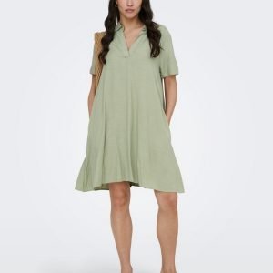 Woman wearing a loose light-green V-neck dress, carrying a woven tote over her shoulder, wearing beige block-heeled sandals.