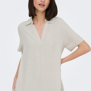 Model wearing a loose, light beige short-sleeve V-neck tunic dress, posing with one hand in a pocket.