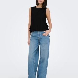 Woman standing in a black sleeveless top and light-wash wide-leg jeans, posing with one hand in her pocket against a pale background.
