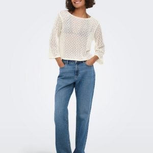Woman wearing a white crochet-knit sweater with blue jeans, standing with hands in pockets and smiling.