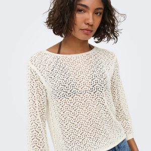 Woman with short curly hair wearing a cream crocheted sweater and blue jeans, posed against a pale background.