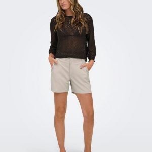 Model wearing a black lace long-sleeve top and beige tailored shorts, hands in pockets, standing in heels against a light gray backdrop.
